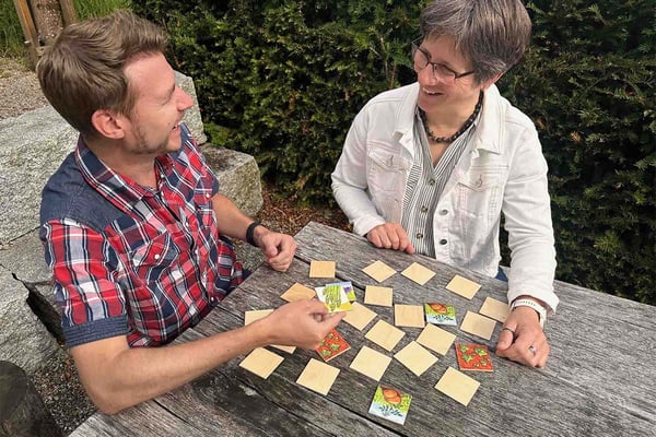 A man and a woman are sitting at a wooden table, laughing and playing Memory.
